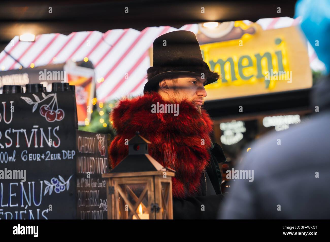 Un venditore distintivo appare con cappello scuro e sciarpa rossa vivace accanto al bancone delle bevande al mercato di Natale per lavoro stagionale, commercio locale. Foto Stock