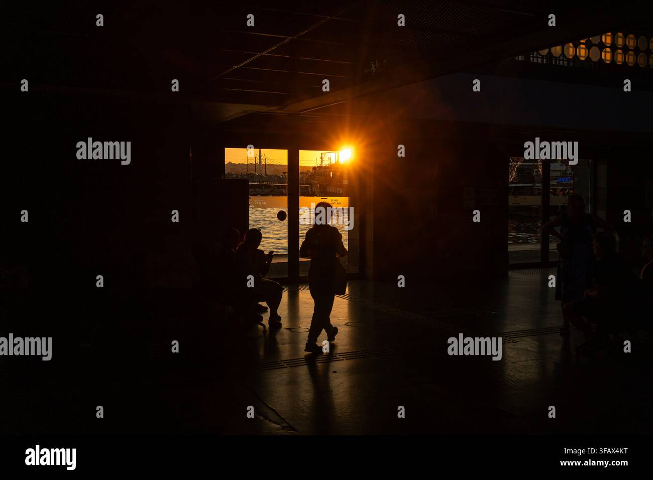 Le persone camminano attraverso uno spazio interno scarsamente illuminato vicino a una grande finestra da cui entra la luce del sole. Foto Stock