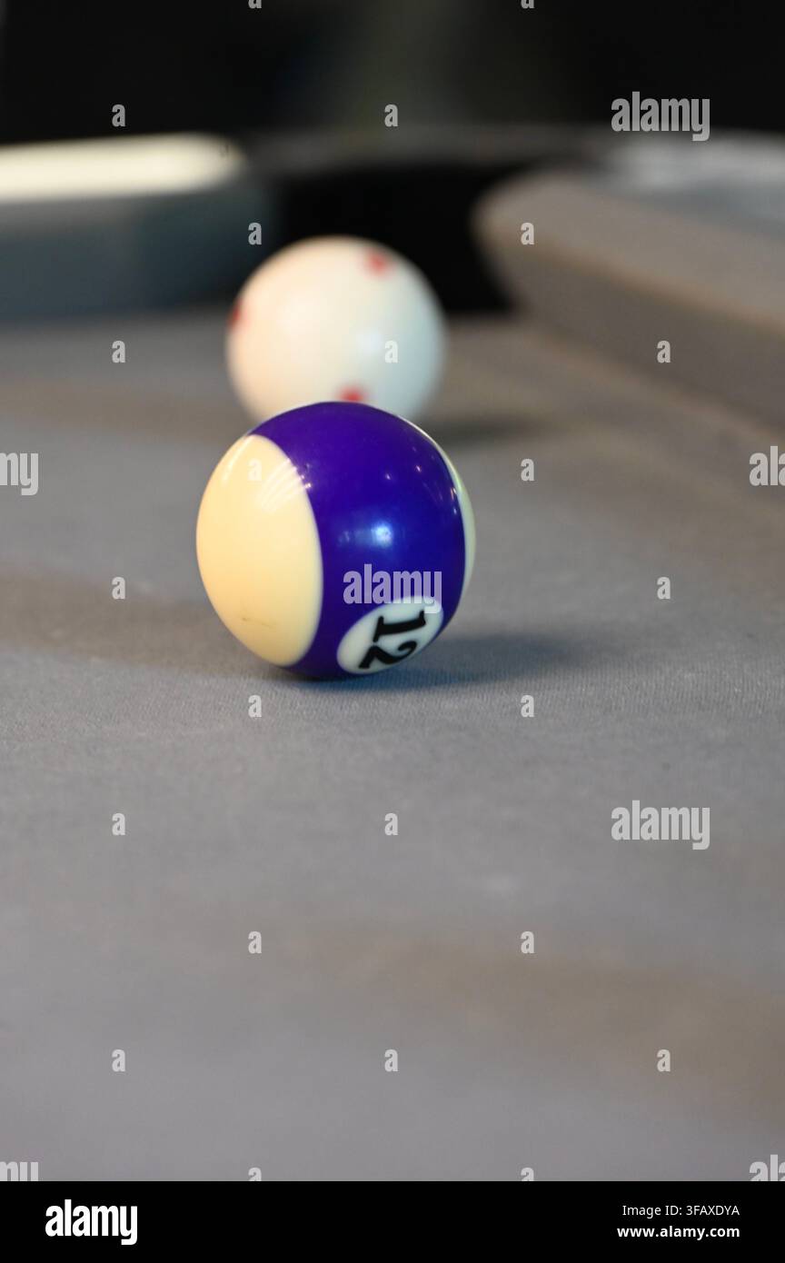 Fotografia verticale in primo piano della palla numerata dodici a strisce viola e bianche a fuoco, con la palla bianca posizionata leggermente dietro vicino al buco di un tavolo da biliardo in feltro grigio. Foto Stock
