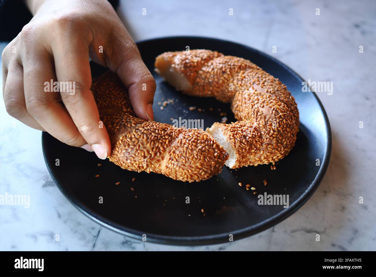 Mano che rompe un panino al sesamo su un piatto nero Foto Stock