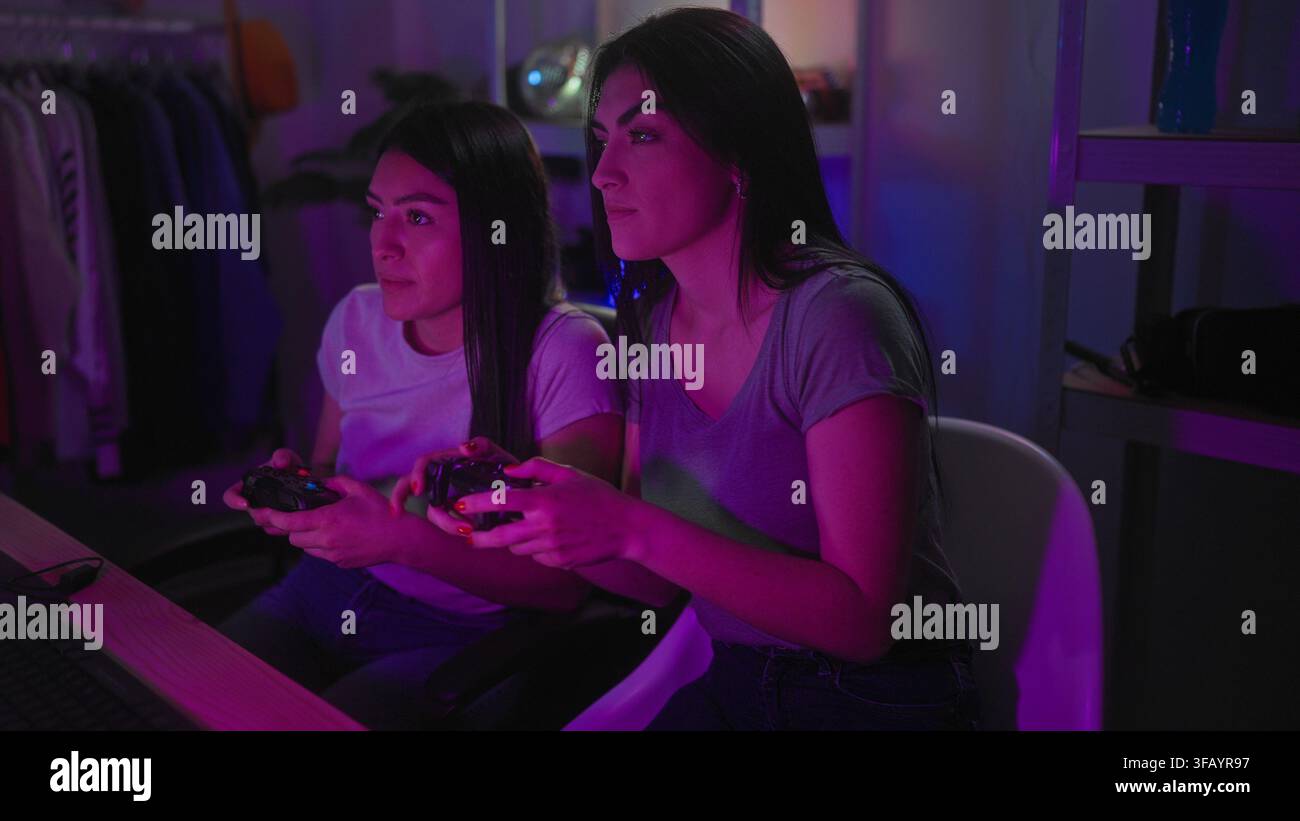 Due donne che tengono i controller del gioco, mani che stringono i joystick, sorridendo e sporgendosi in avanti mentre giocano in uno studio con scaffali neon e lampada; eccit Foto Stock