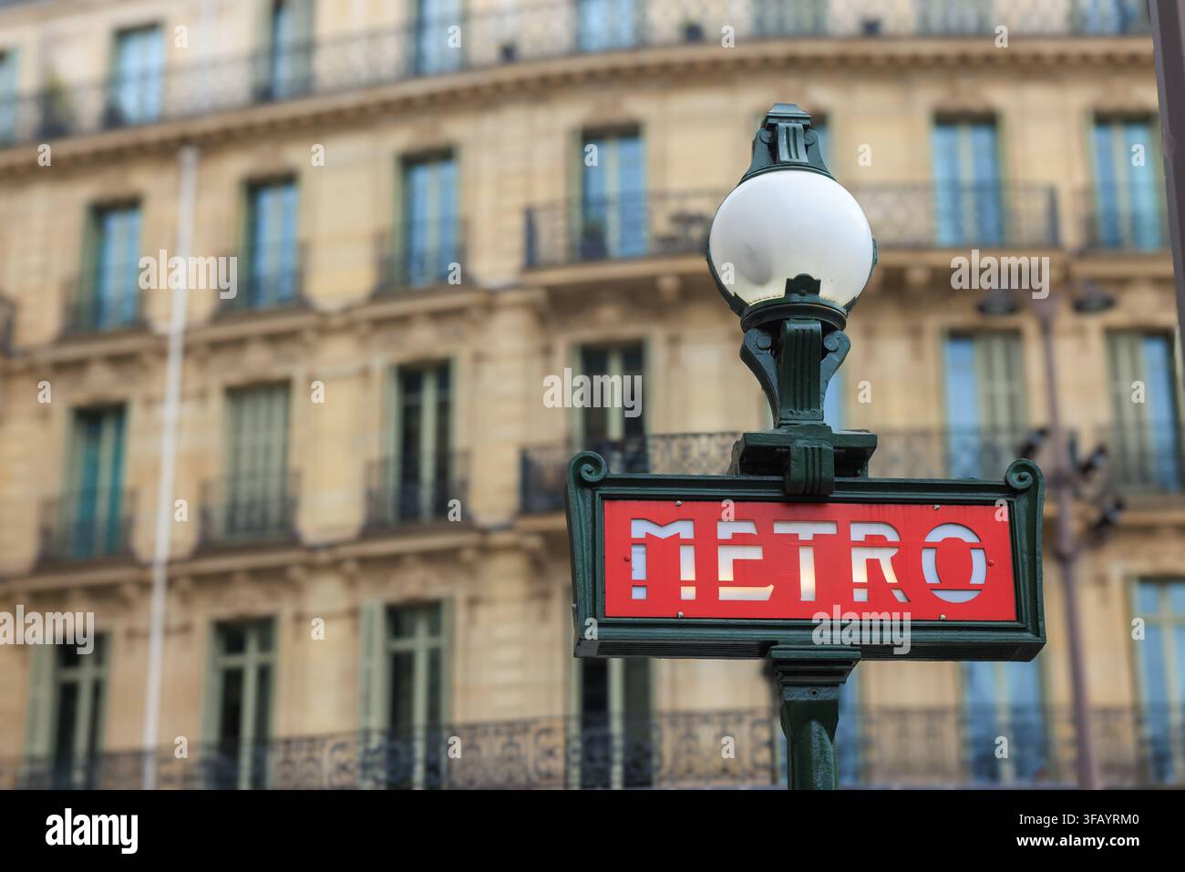Insegna classica rossa "Metro" con un palo verde ornato e una luce a globo davanti a un edificio residenziale in stile Haussmann sfocato, Parigi, Francia. Foto Stock