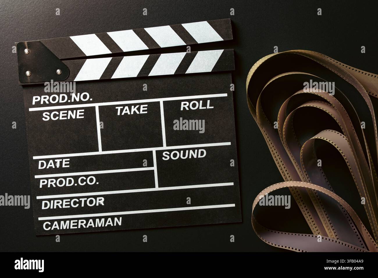 Claquette cinematografica con striscia di film su sfondo nero, primo piano. Vista dall'alto della classica claquetta nera accanto a una striscia di film vintage srotolata Foto Stock