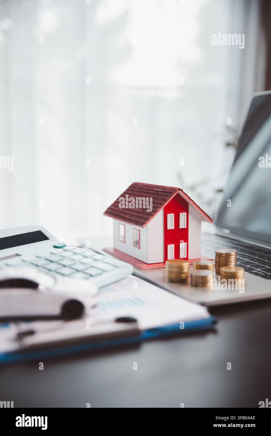 Modello di casa su un laptop accanto a calcolatrice, monete, documenti e auto rappresenta la pianificazione del mutuo digitale, il budget online, il finanziamento immobiliare, veicolo Foto Stock