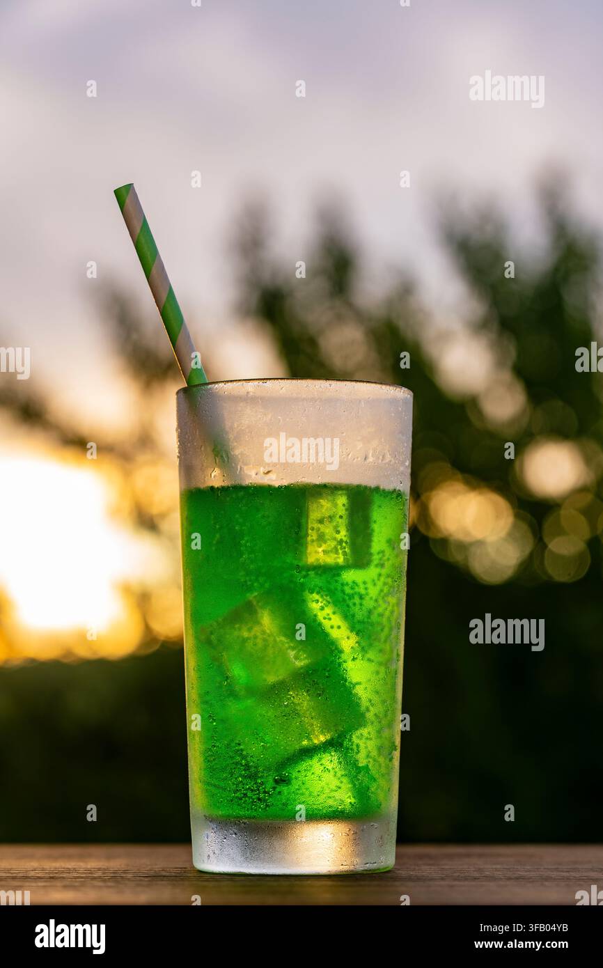 Bevanda gassata verde fredda con ghiaccio su sfondo al tramonto. Bicchiere alto rinfrescante di un cocktail gassato verde vivace con cubetti di ghiaccio e cannuccia a strisce, piazza Foto Stock