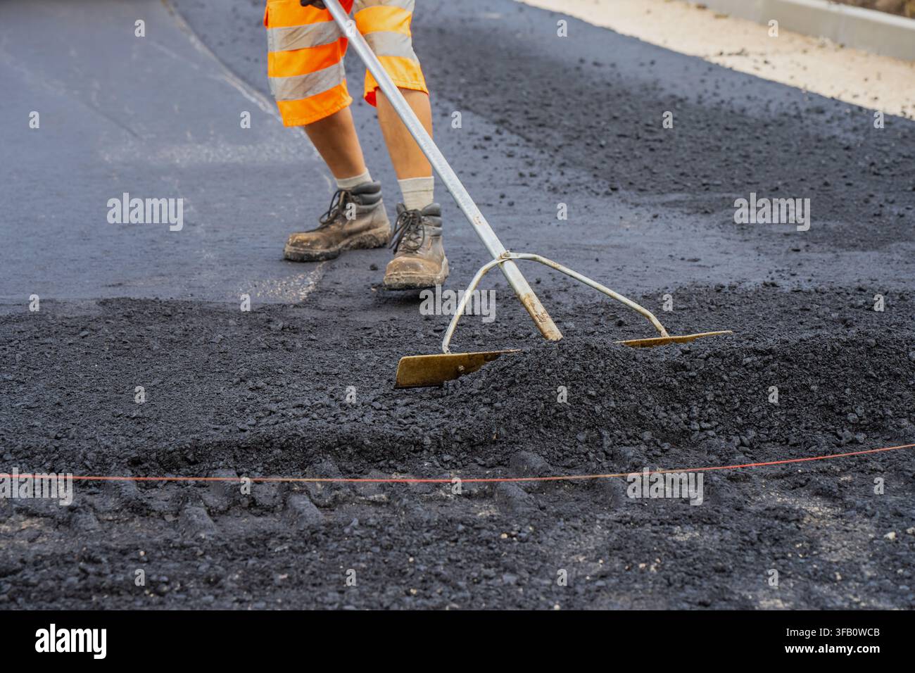 Lavoratore edile che utilizza una cazzuola per asfalto a manico lungo per livellare manualmente lo strato superficiale di asfalto caldo durante i lavori di pavimentazione stradale e ingegneria civile Foto Stock