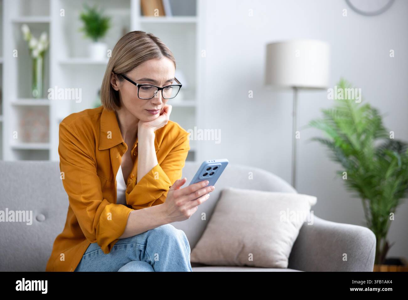 Donna con occhiali seduta su un comodo divano a casa, immersa nel suo smartphone, connettendosi con le persone, navigando sui social media o lavorando da remoto Foto Stock