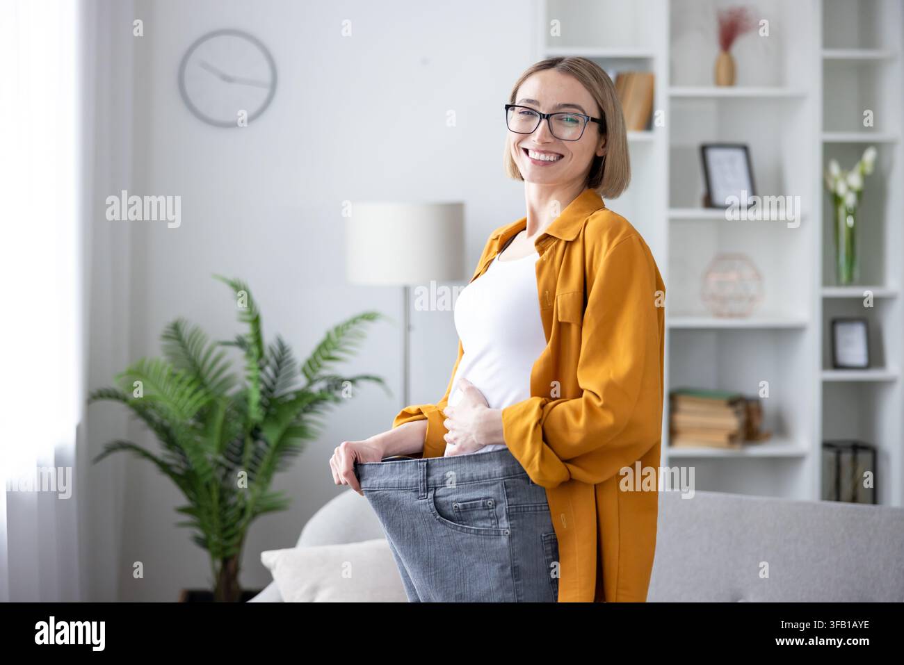 Donna felice che tiene in alto i suoi jeans larghi, celebrando una perdita di peso riuscita e una trasformazione dello stile di vita sano, dimostrando progressi nel suo percorso di fitness a casa Foto Stock