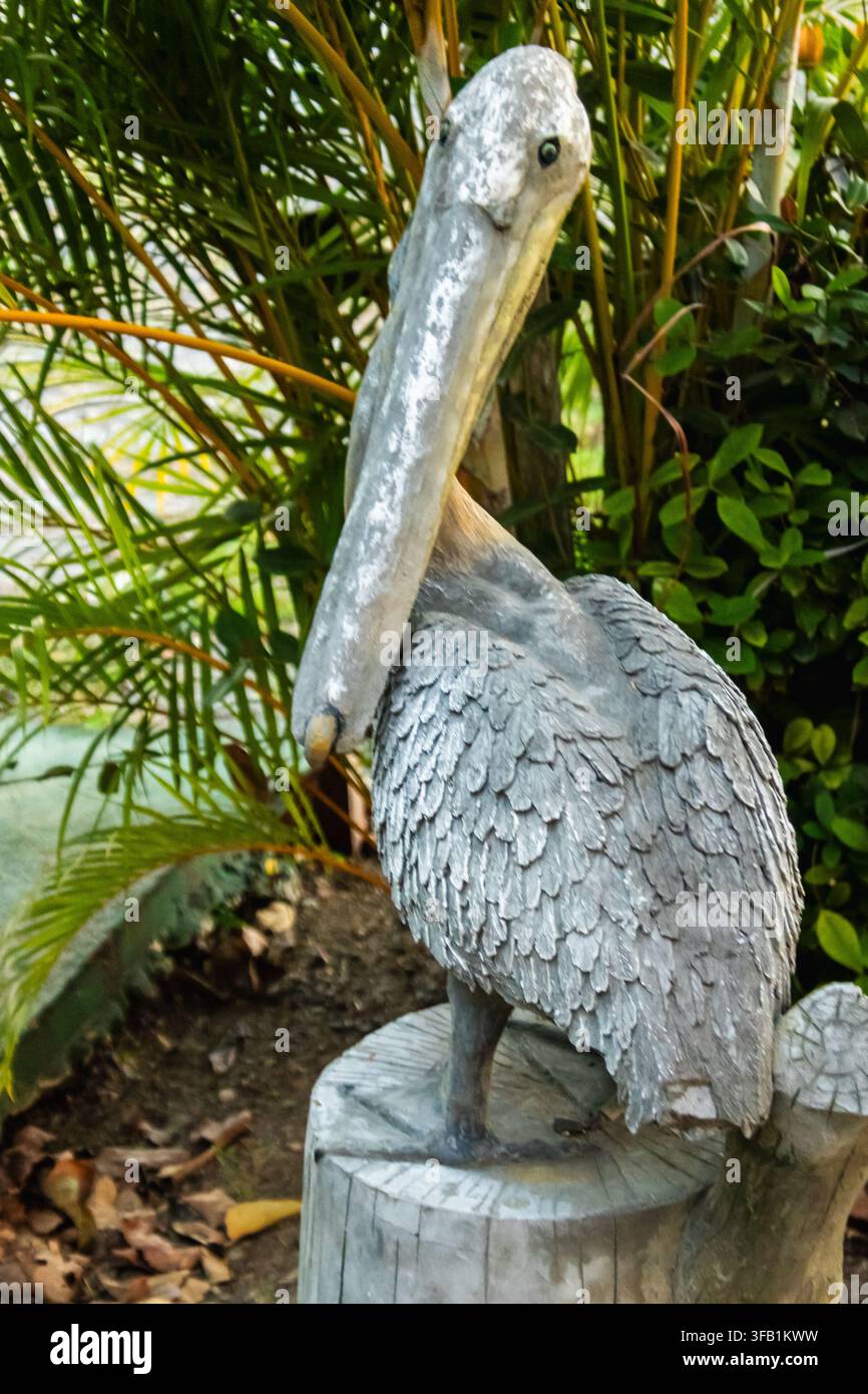Statua da giardino di un pellicano grigio consumata dalle intemperie, in piedi su un ceppo d’albero tra lussureggianti foglie di palme tropicali e piante verdi all’aperto Foto Stock