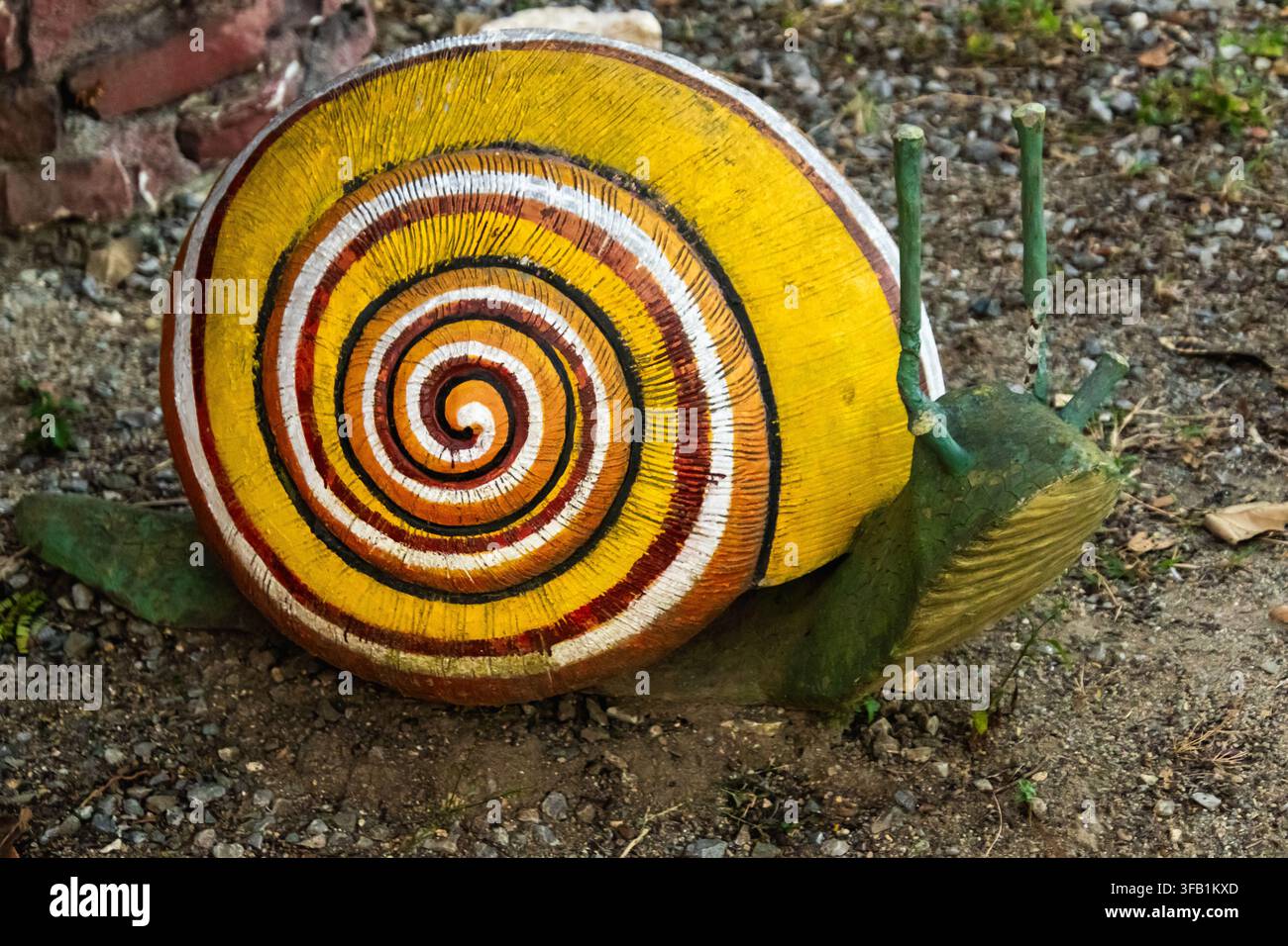 Scultura di lumaca colorata fatta a mano con una conchiglia a spirale giallo brillante, posata sul terreno in un ambiente rustico da giardino. Foto Stock