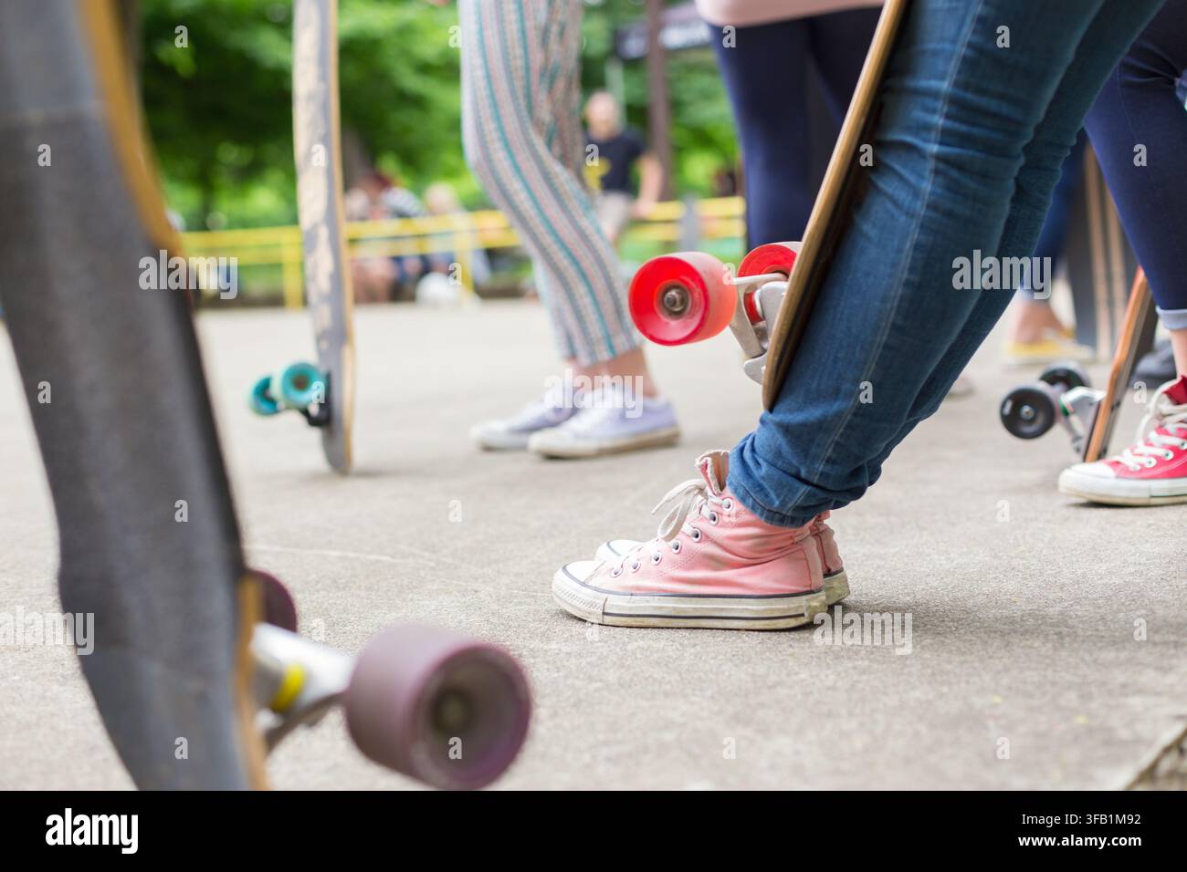 Primo piano ad angolazione bassa di adolescenti in piedi sul marciapiede con i loro longboard e skateboard, che mostrano scarpe da ginnastica rosa e ruote colorate dello skateboard in un parco skate all'aperto Foto Stock