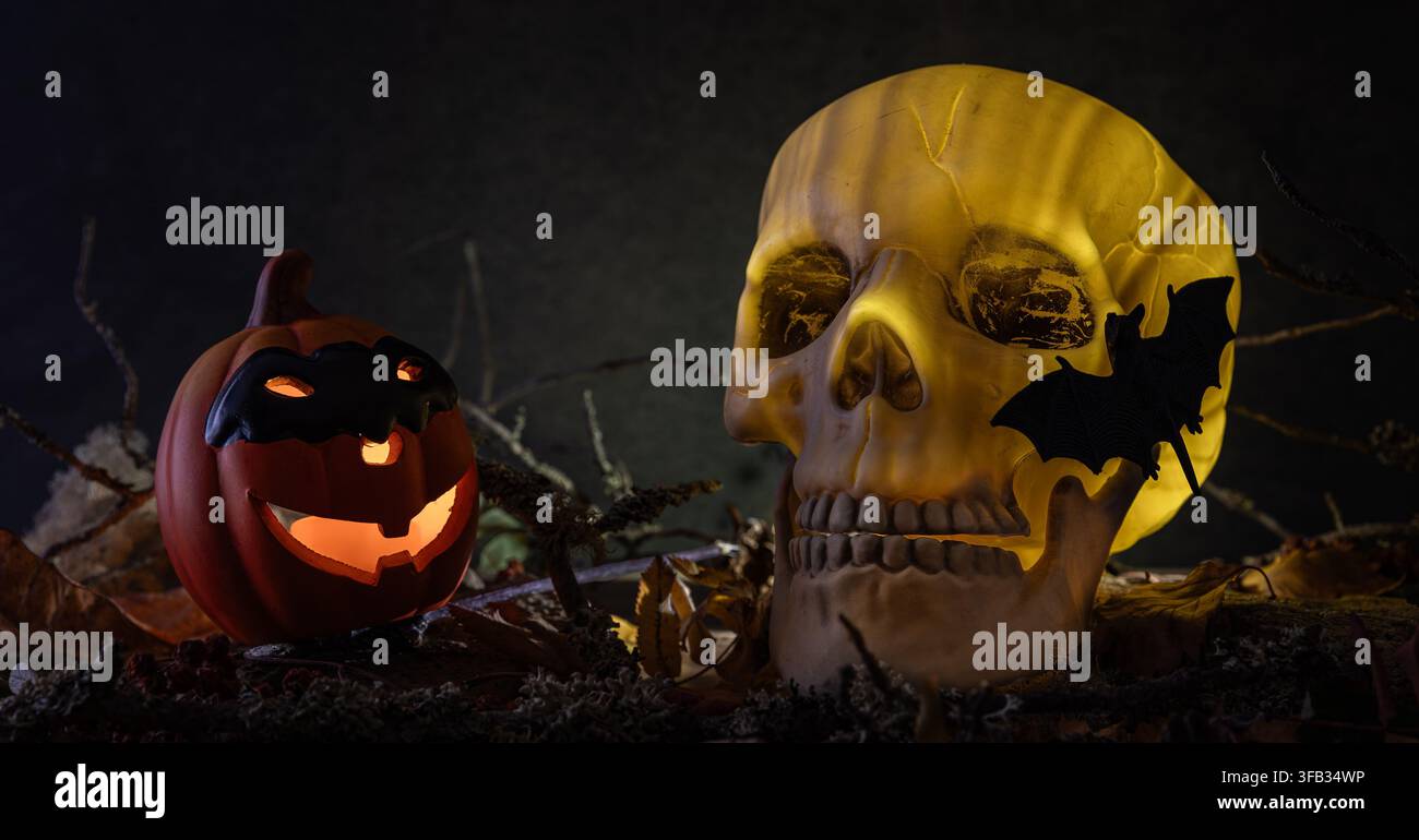 Teschio umano luminoso e inquietante con un pipistrello sopra e una lanterna di zucca sorridente posizionata tra foglie e rami appassiti su uno sfondo scuro. Foto Stock