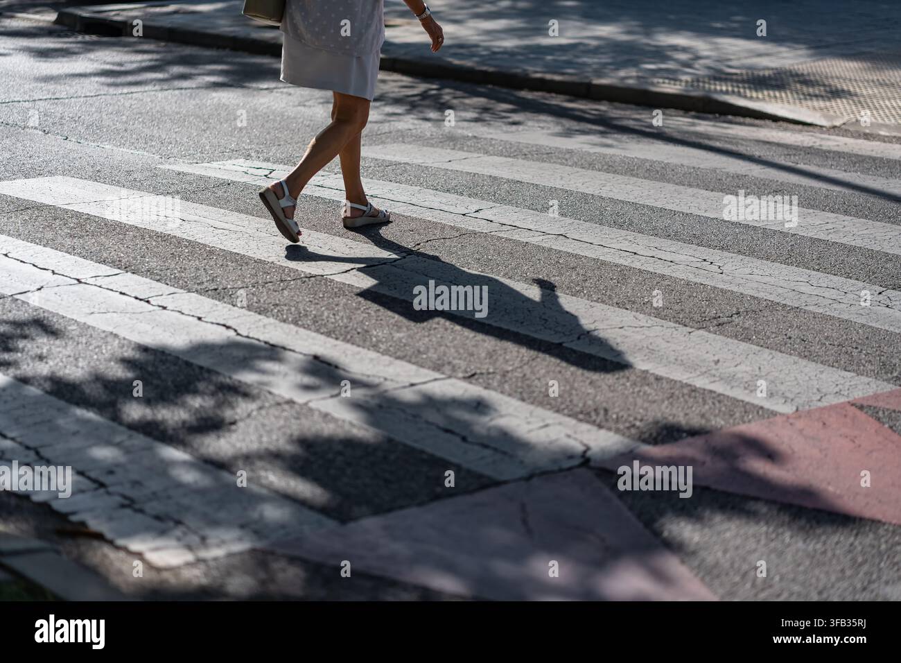 Donna che attraversa un passaggio pedonale a zebre con lunghe ombre su una strada urbana luminosa e soleggiata Foto Stock