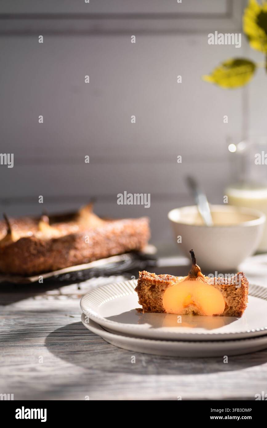 Prima piano di una fetta di pan di spagna alle pere sormontata da una pera poché su un piatto bianco, con il resto della torta, una ciotola, un cucchiaio e fiori gialli in un su Foto Stock