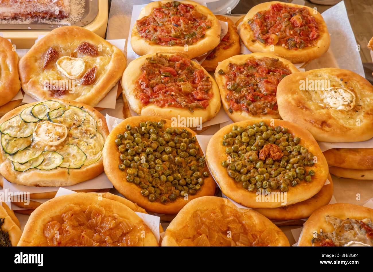 Una selezione di mini pizze rotonde è disposta su un tavolo in un mercato alimentare. Queste pizze presentano condimenti come peperoni rossi, piselli verdi, cipolle caramellizzate Foto Stock