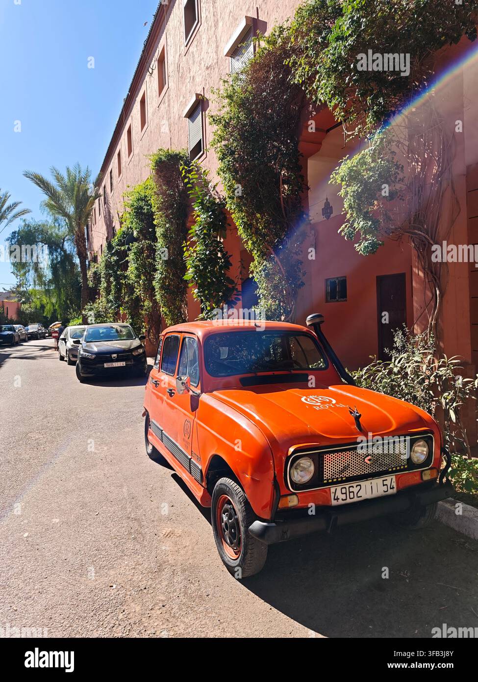 Renault 4 arancione vintage parcheggiato su una strada a Marrakesh, in Marocco, con palme, vegetazione lussureggiante e edifici in stile marocchino rosa. - Immagine stock catturata con smartphone