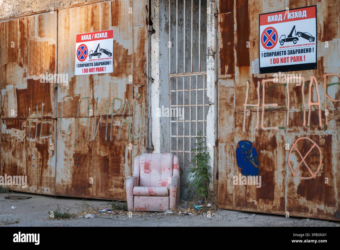 Una poltrona abbandonata e logorata è posizionata sulla strada davanti a porte metalliche industriali pesantemente arrugginite, segnate da graffiti sbiaditi e da una scritta "No Parking" Foto Stock