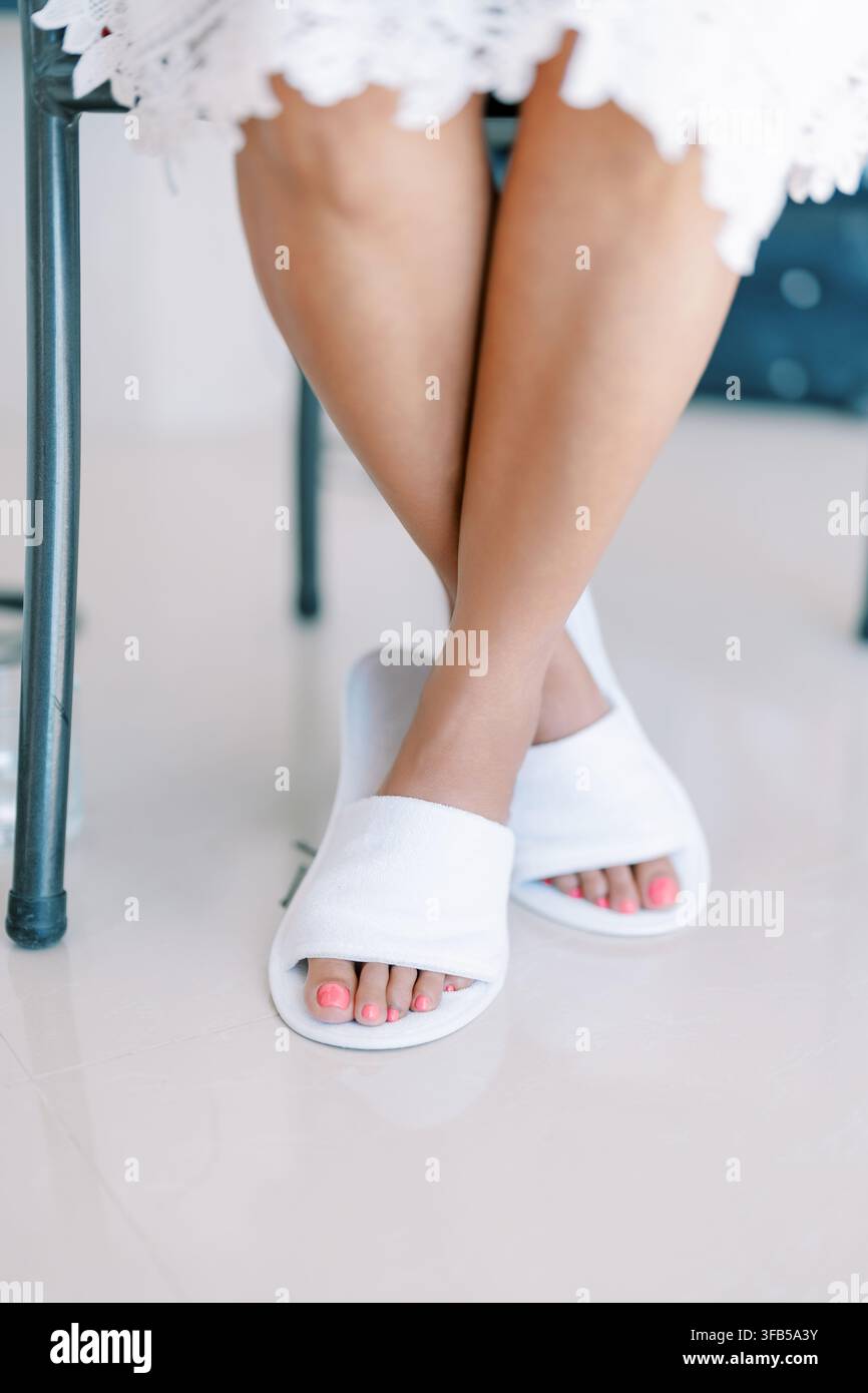 Gambe incrociate con pantofole bianche e unghie dei piedi rosa Foto Stock