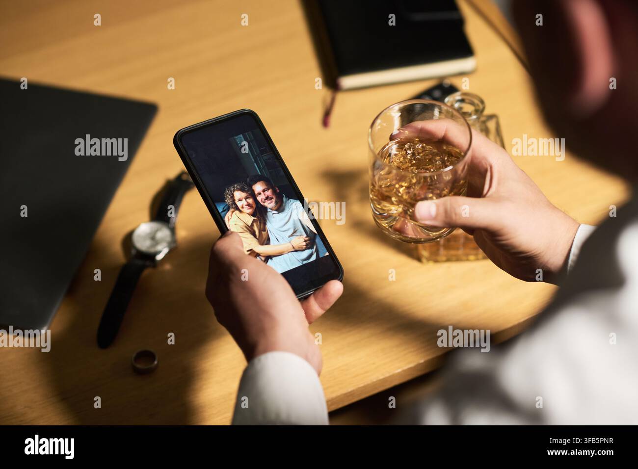 Uomo depresso che tiene un bicchiere di alcol e guarda una foto sul suo smartphone Foto Stock