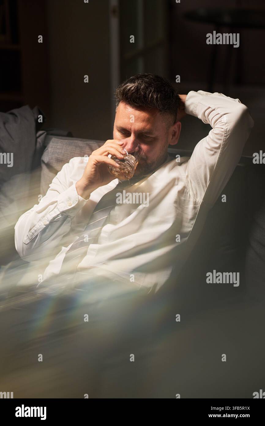 Uomo d'affari di mezza età che beve whisky sul divano dopo una dura giornata di lavoro Foto Stock