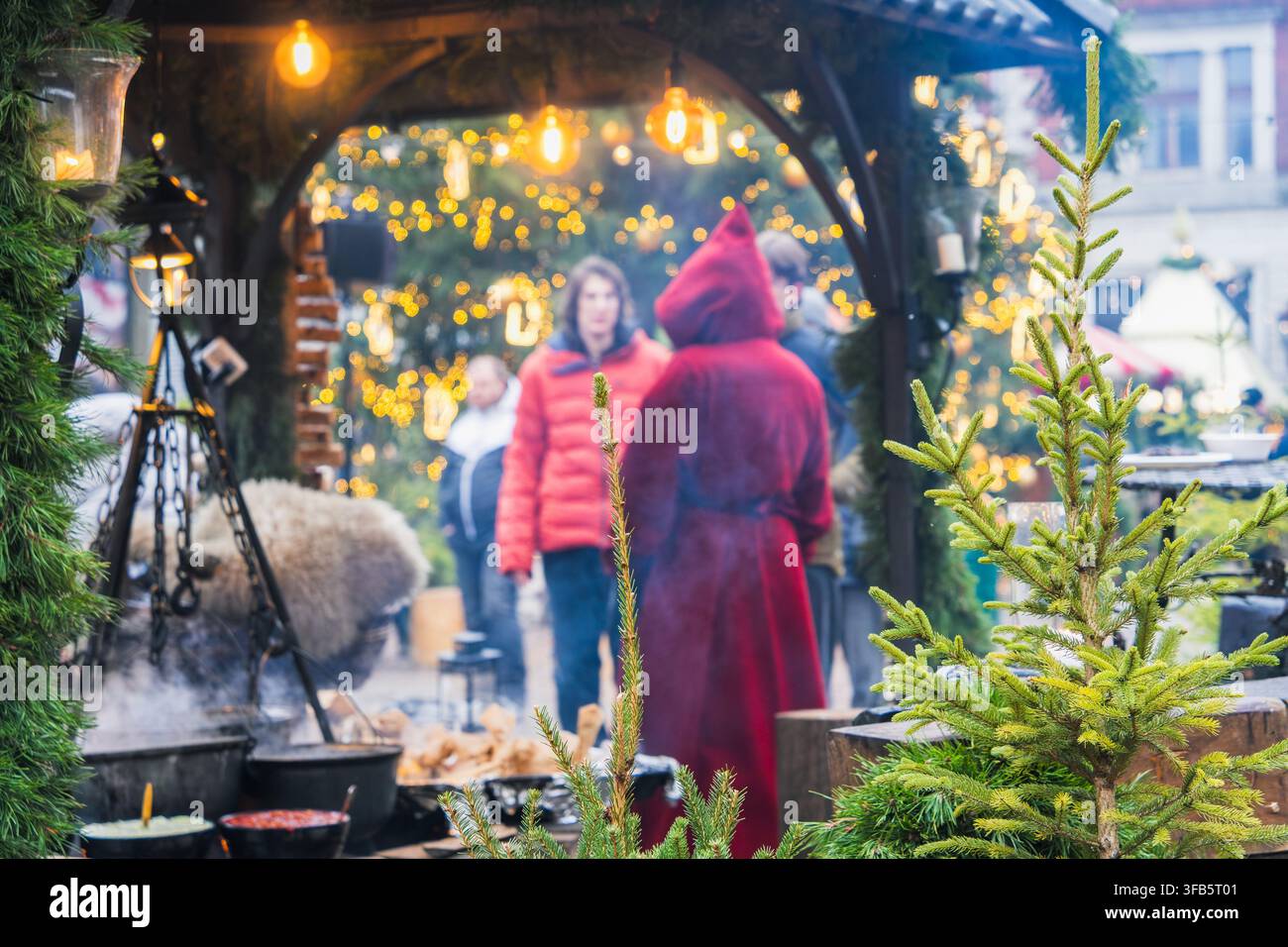 Vista stratificata delle feste guarda attraverso un arco sempre verde verso i visitatori, gli abeti e gli stand luminosi del mercato di Natale di Riga per un viaggio atmosferico Foto Stock