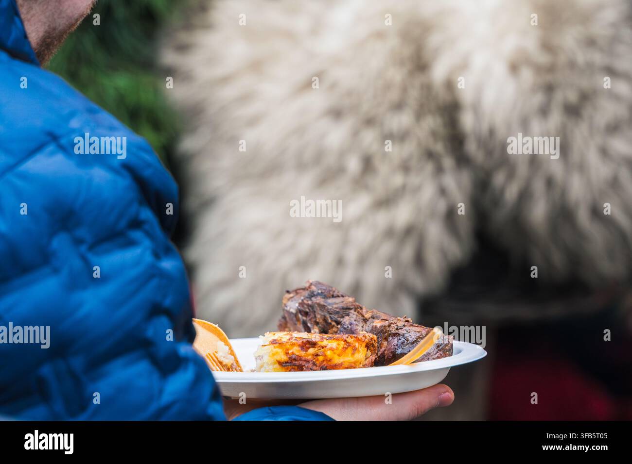 Un pasto caldo da strada viene trasportato attraverso il mercato natalizio di Riga mentre un visitatore tiene un piatto di cibo grigliato contro le morbide texture invernali della cucina locale Foto Stock