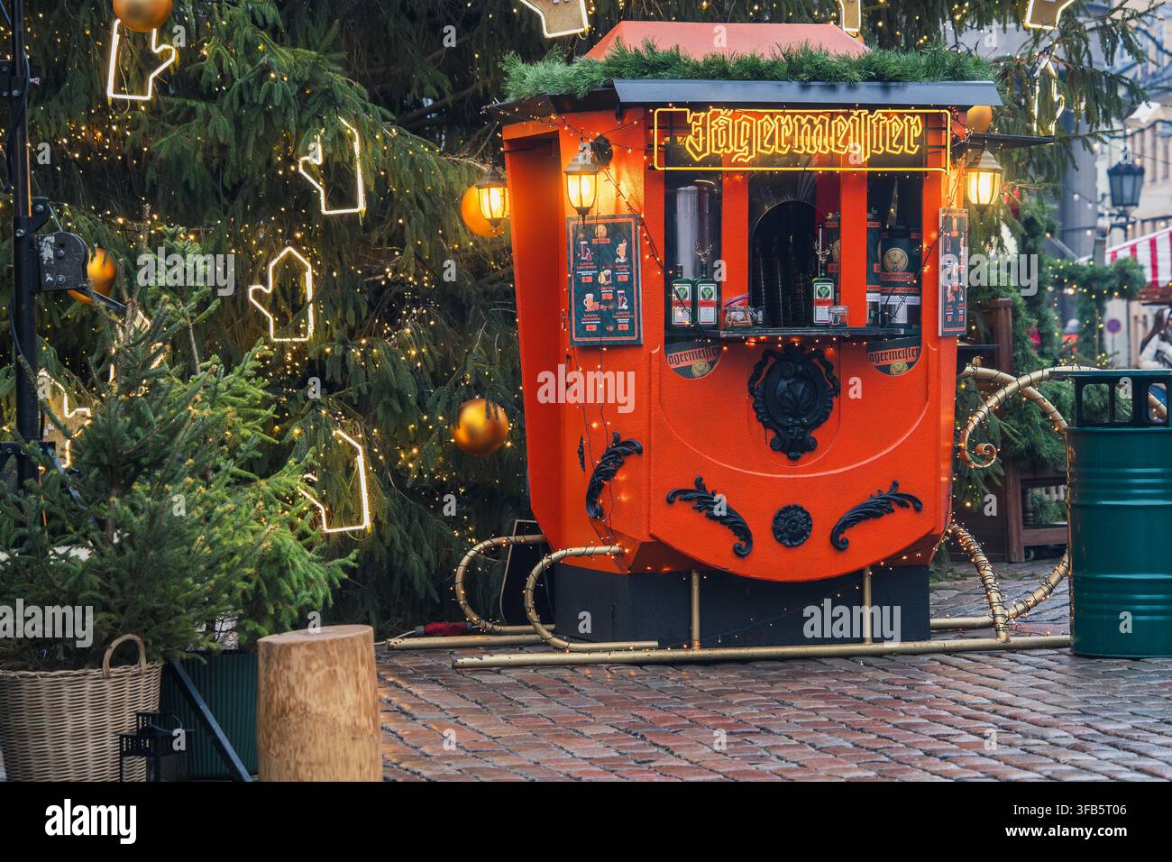 Chiosco di bevande colorato risplende di arancione tra i rami degli abeti decorati al mercato di Natale di Riga, presentando un servizio di bevande calde per l'ospitalità invernale, touri Foto Stock