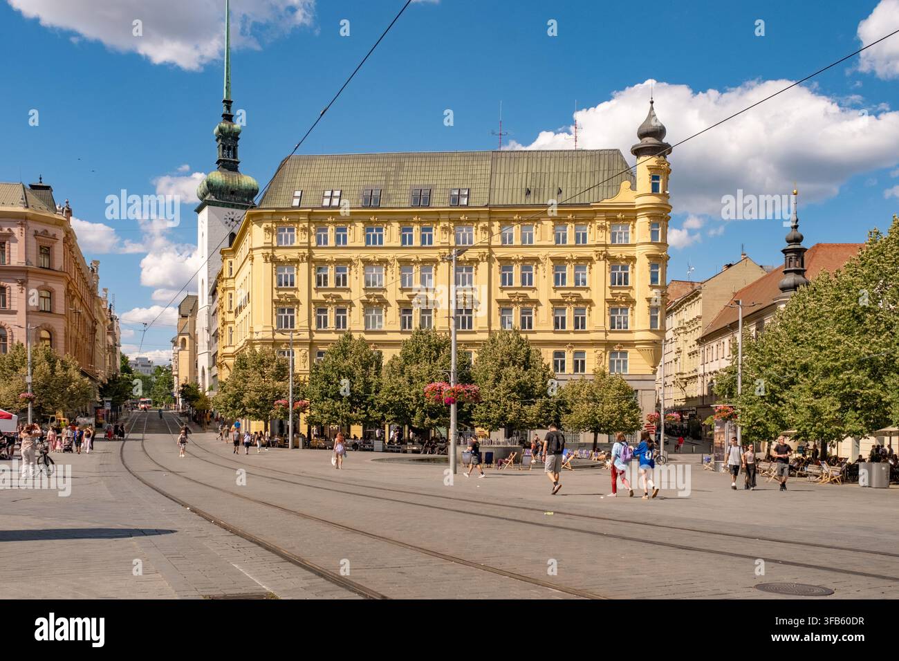 Piazza urbana soleggiata con edificio storico giallo ornato, guglia della chiesa, binari del tram e vivace scena pedonale sotto un cielo blu terso Foto Stock