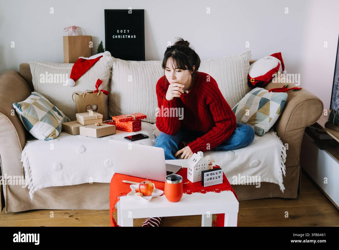 Donna in maglione di lana lavora su un laptop sul divano accanto a pacchi incartati e un cartello HOLLY JOLLY. Lavoro da remoto dicembre, ufficio domestico con laptop Natale Foto Stock