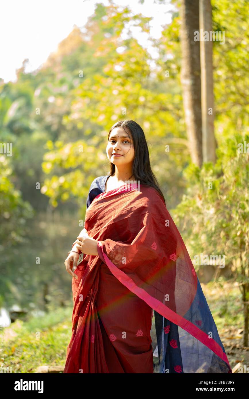 Una donna con un saree rosso si trova all'aperto in un ambiente naturale con vegetazione e luce solare che filtra attraverso gli alberi. Foto Stock
