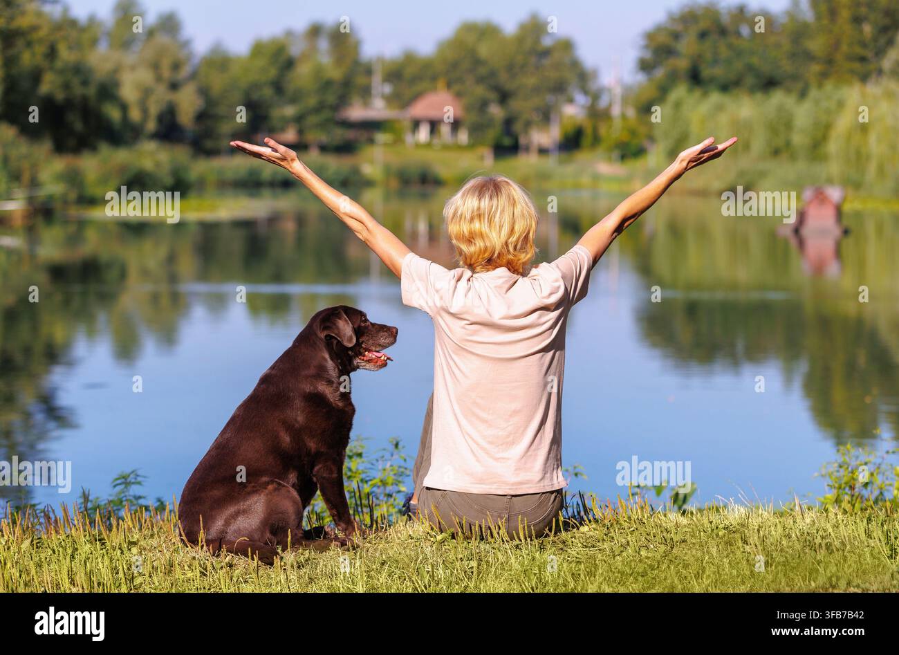 Donne che godono di una vista panoramica sul lago con il loro Labrador Retriever cioccolato nel parco cittadino, alzando le braccia con gioia, tranquillo svago all'aperto, app natura Foto Stock
