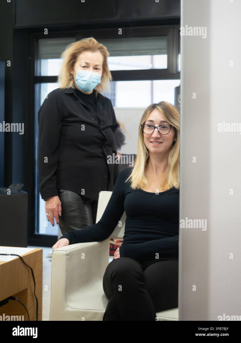 Due donne professionali in un ambiente d'ufficio, una con una mascherina sul viso in piedi dietro una collega seduta che sorride Foto Stock