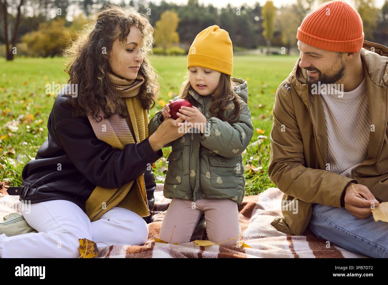Genitori e figlia condividono una mela durante un accogliente picnic autunnale nel parco, con sorrisi Foto Stock