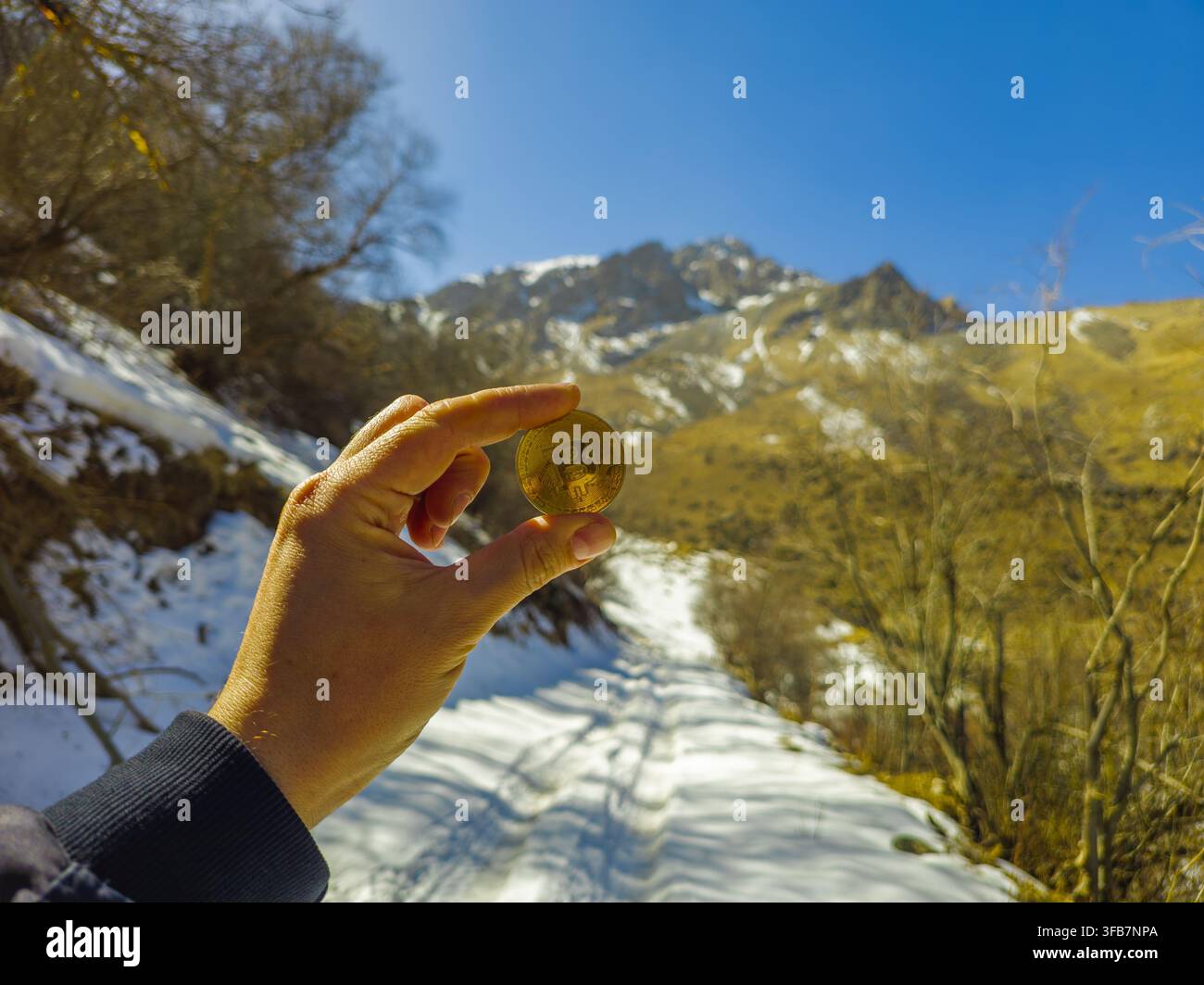 Una mano tiene una moneta d'oro della criptovaluta contro uno sfondo di montagne innevate nel Kirghizistan. La scena contrasta la moneta digitale con la bellezza naturale un Foto Stock
