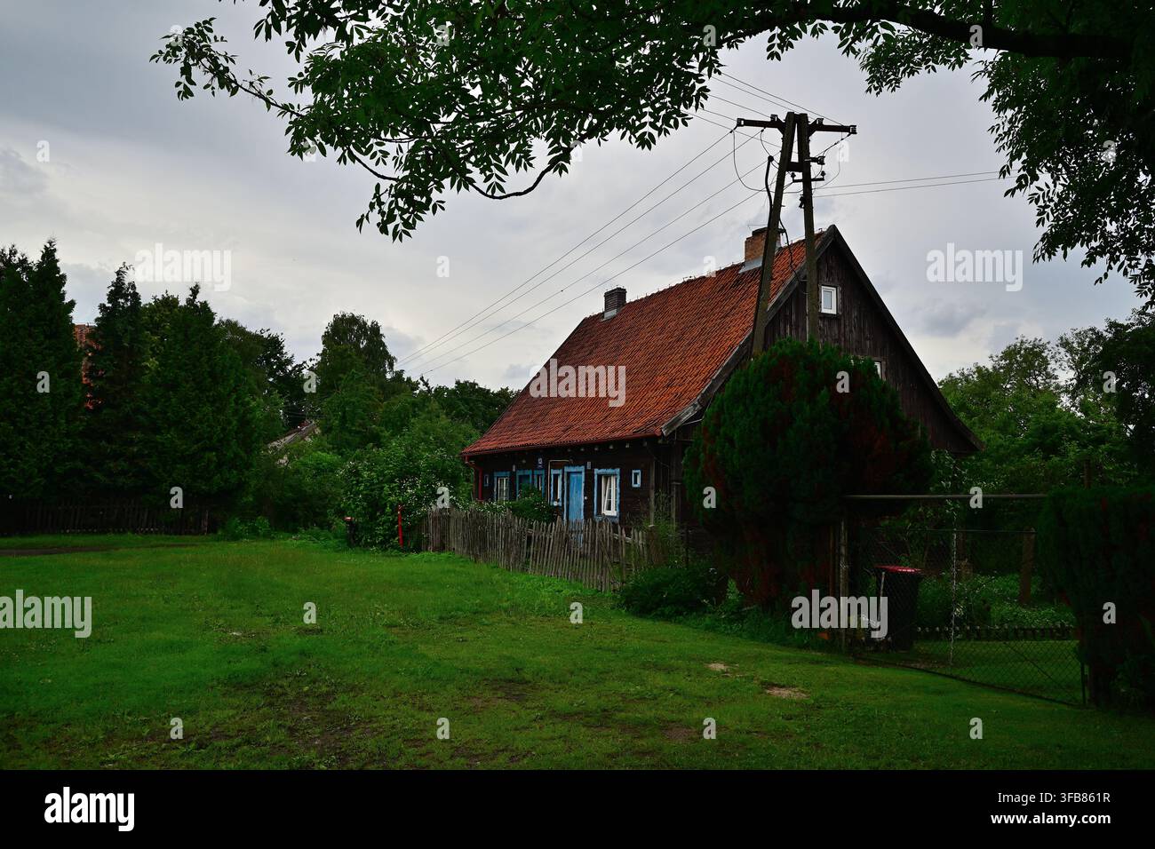 Chata Mazurska (Cottage Masuriano) Cottage rurale rustico con tetto di tegole rosse e giardino recintato sotto un cielo cupo nel paesaggio del villaggio - Popielno in Masuria Foto Stock