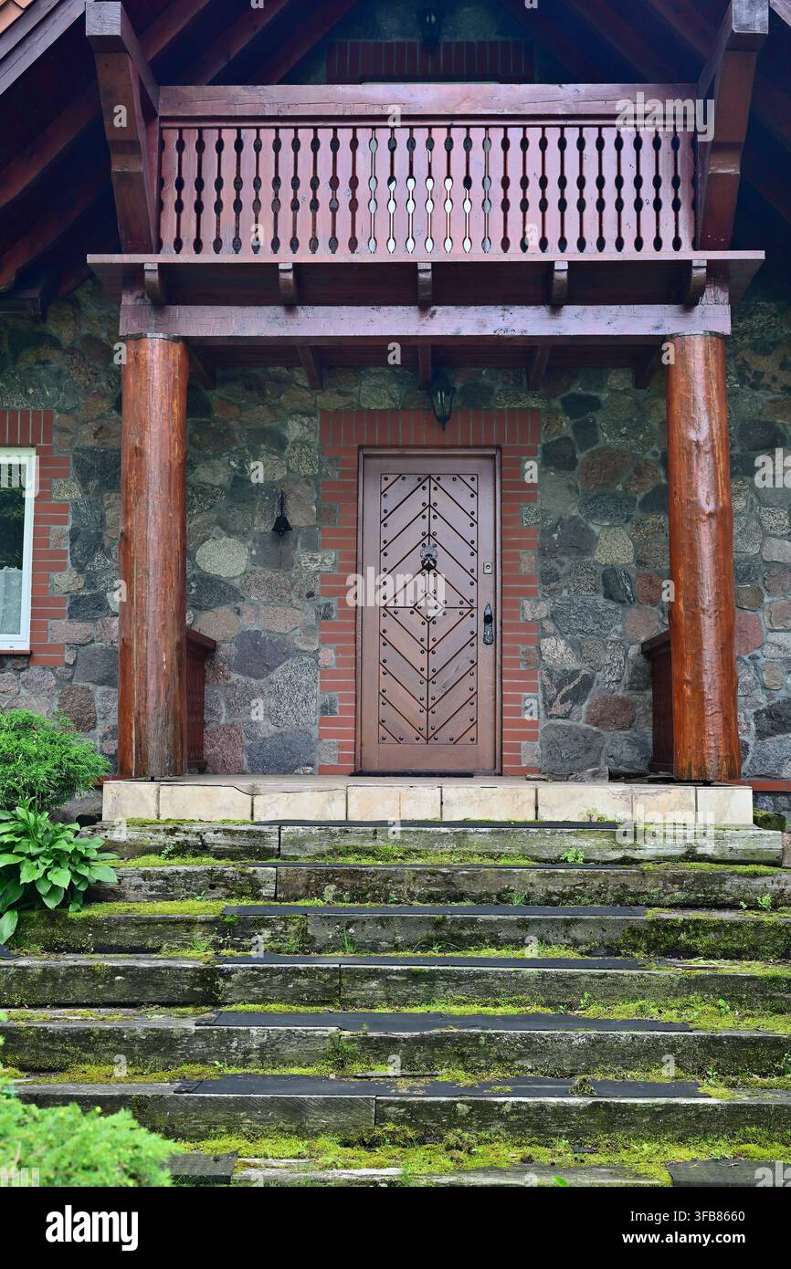 Ingresso di casa in pietra rustica con porta in legno intagliata, gradini muschiosi e portico con balcone Foto Stock