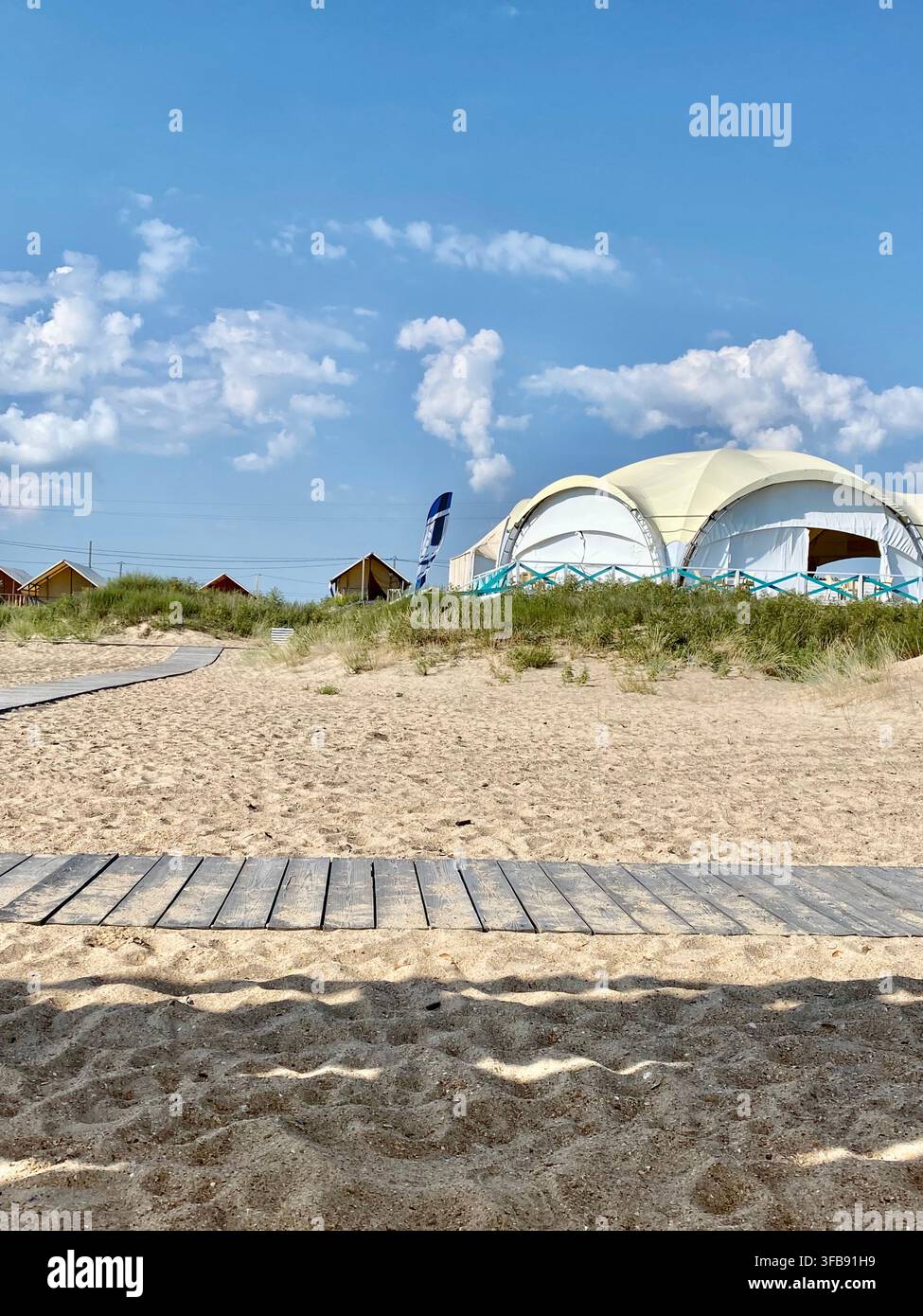Villaggio glamping costiero con strutture in tela e sentieri in legno sotto il cielo estivo - Immagine stock catturata con smartphone