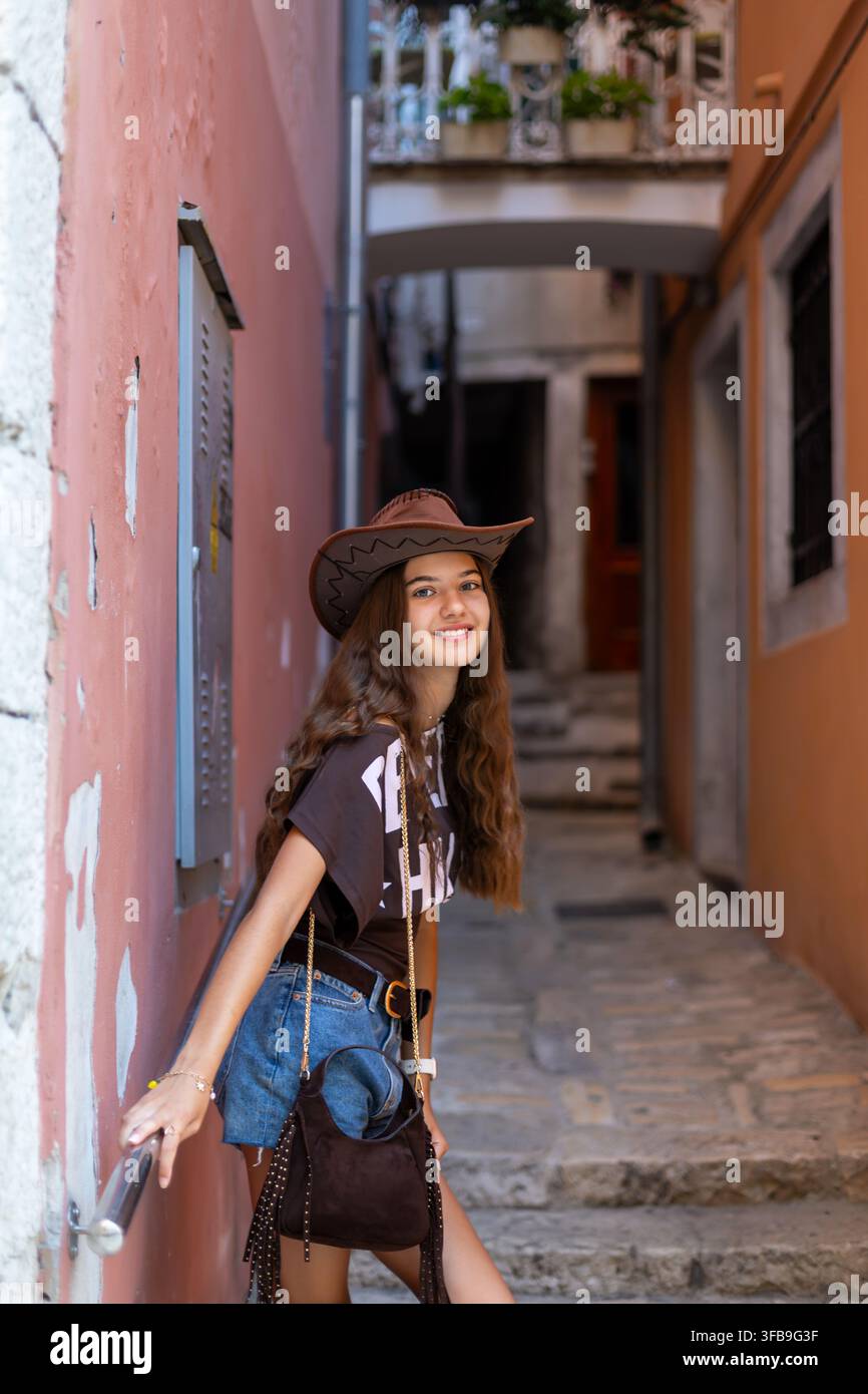 Giovane donna con cappello da cowboy su una stretta scala in pietra. Giovane donna con un cappello da cowboy marrone e pantaloncini di jeans sorride su una stretta scala in pietra tra Foto Stock