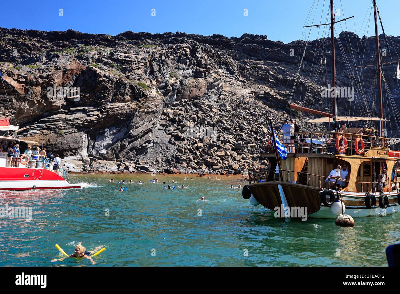 Santorini, Grecia – 4 agosto 2026: Turisti nuotano in acque terapeutiche ricche di minerali vicino a barche in una baia vulcanica Foto Stock