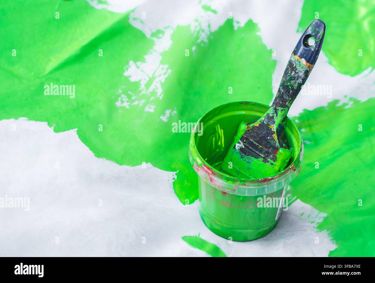 Piccolo secchio pieno di vernice verde vivace e un pennello che vi riposa, su una superficie bianca con schizzi di vernice verde Foto Stock