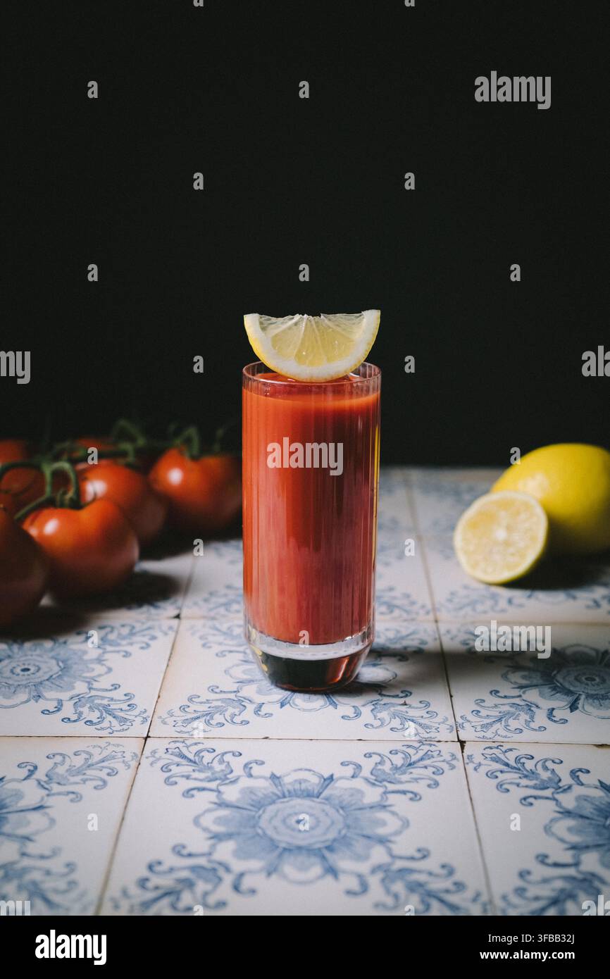 Un fresco cocktail Bloody Mary guarnito con limone su piastrelle vintage blu, circondato da pomodori freschi su uno sfondo scuro Foto Stock