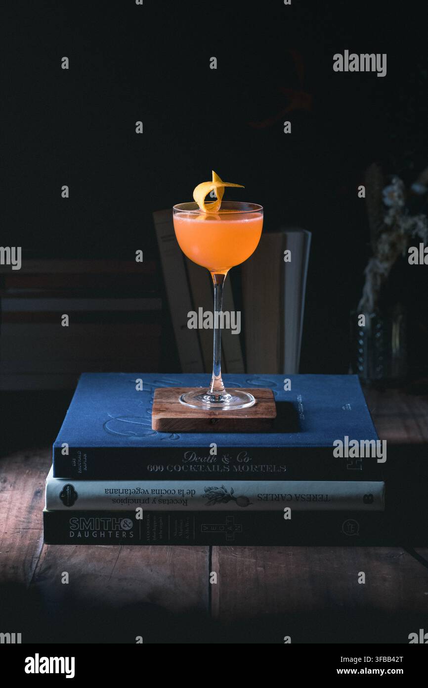 Cocktail artigianale arancione in un bicchiere da coupe su una pila di libri di cocktail in una stanza buia e suggestiva, fotografia di un barista casalingo con spazio per il testo Foto Stock