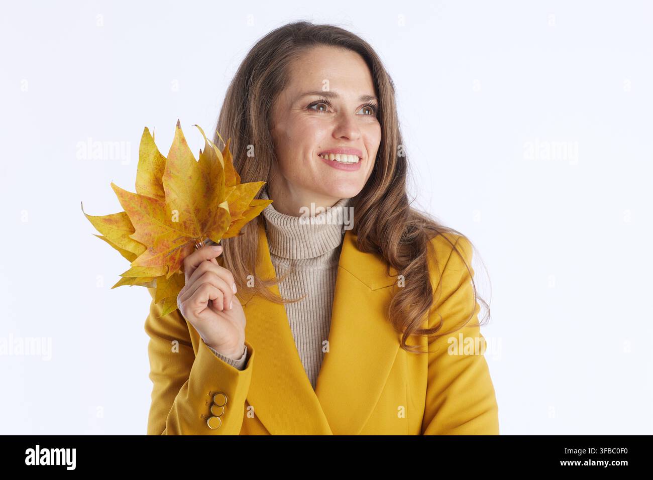 Una donna sorridente con capelli castani in un blazer giallo e una maglia a collo alto beige tiene foglie d'acero gialle e arancioni. Guarda di lato, evocando l'autunno Foto Stock
