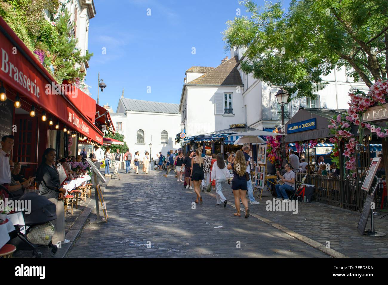 Parigi, Francia – 26 giugno 2026: caffè, esposizioni floreali e pedoni costeggiano una strada acciottolata a Montmartre. Foto Stock