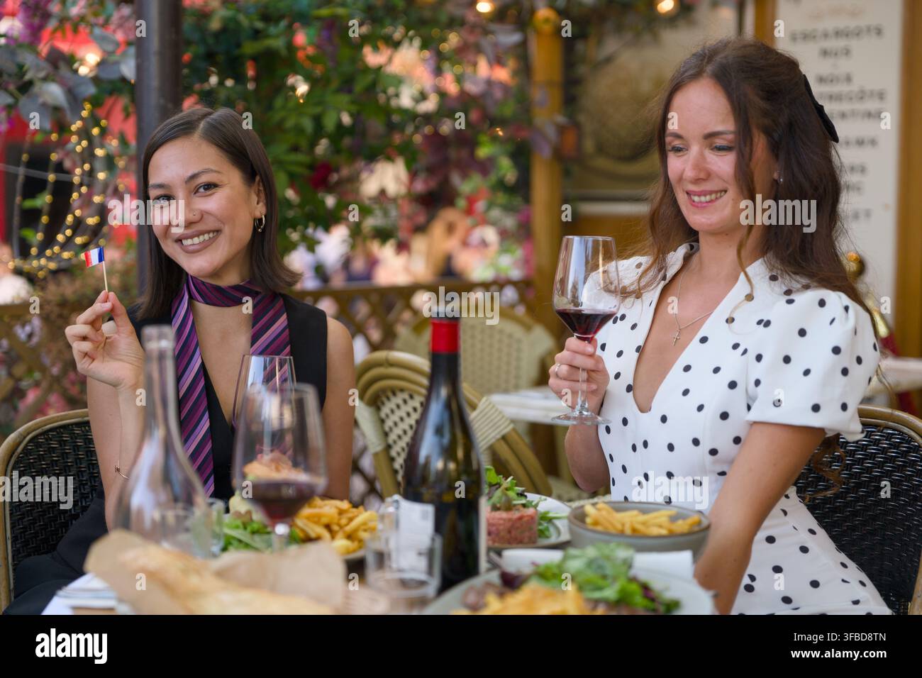 Due donne sorridenti cenano con vino rosso in un ristorante parigino; una tiene una piccola bandiera francese. Foto Stock