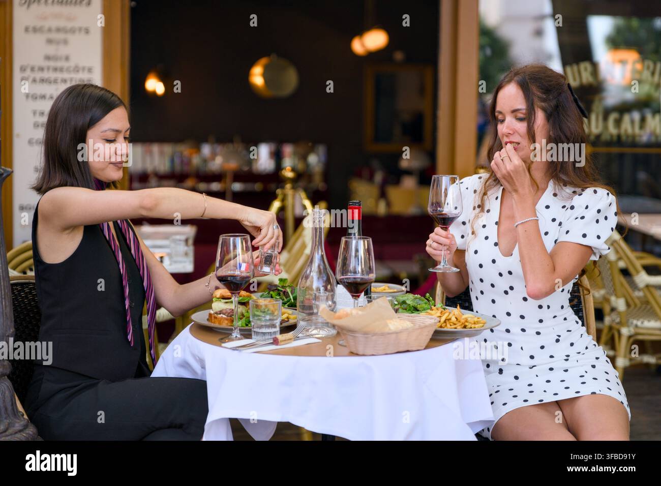 Due donne cenano con vino rosso, patatine fritte e insalata a un tavolo all'aperto di un ristorante a Parigi, Francia. Foto Stock