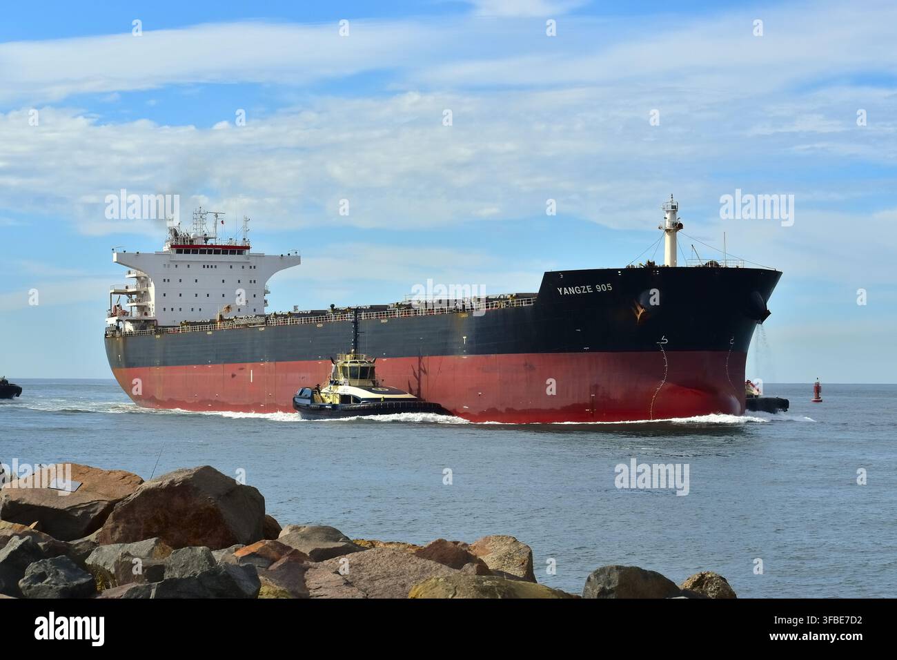 Una nave da carico nera e rossa si muove attraverso acque costiere calme sotto un cielo parzialmente nuvoloso, assistita da una chiatta posizionata vicino alla sua prua. Foto Stock