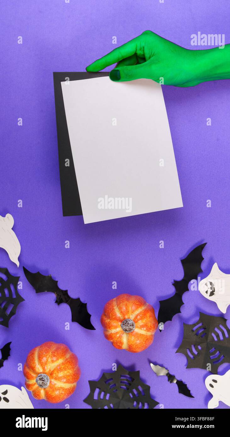 Mano di strega verde che tiene un modello di carta bianca su uno sfondo viola di Halloween Foto Stock