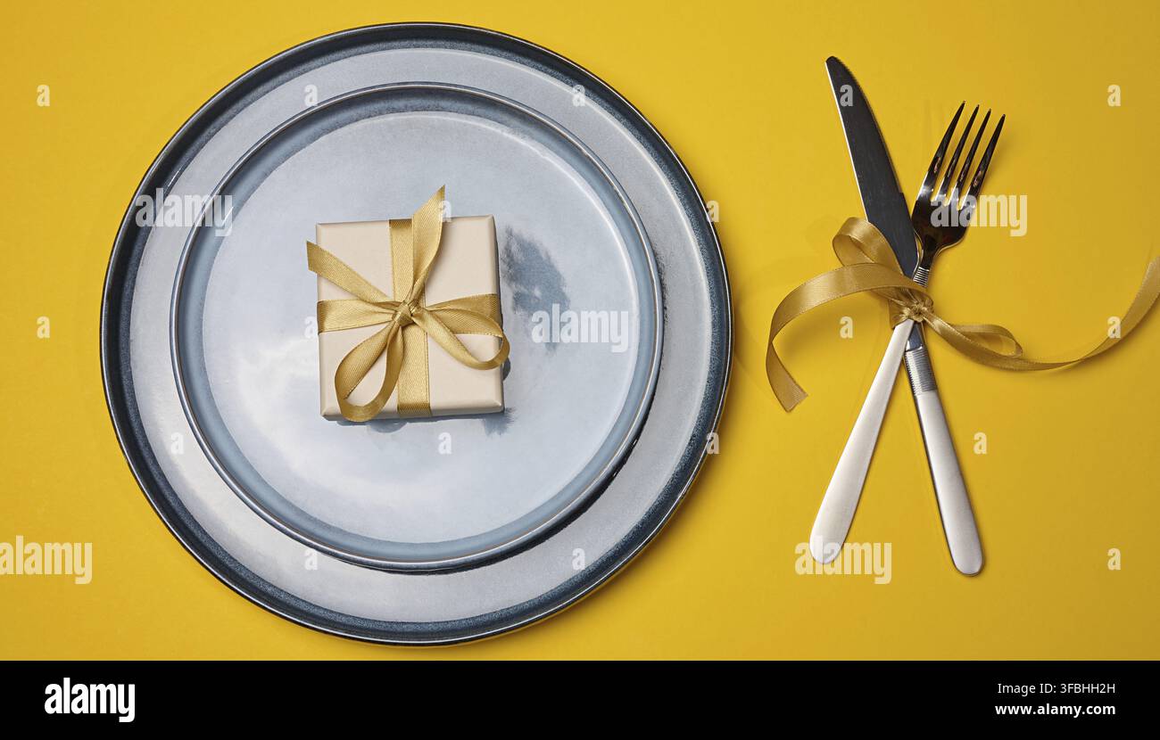 Una tavola festosamente apparecchiata su uno sfondo giallo con un piatto grigio e una scatola regalo. A destra ci sono un coltello e una forchetta, che con uovo Foto Stock