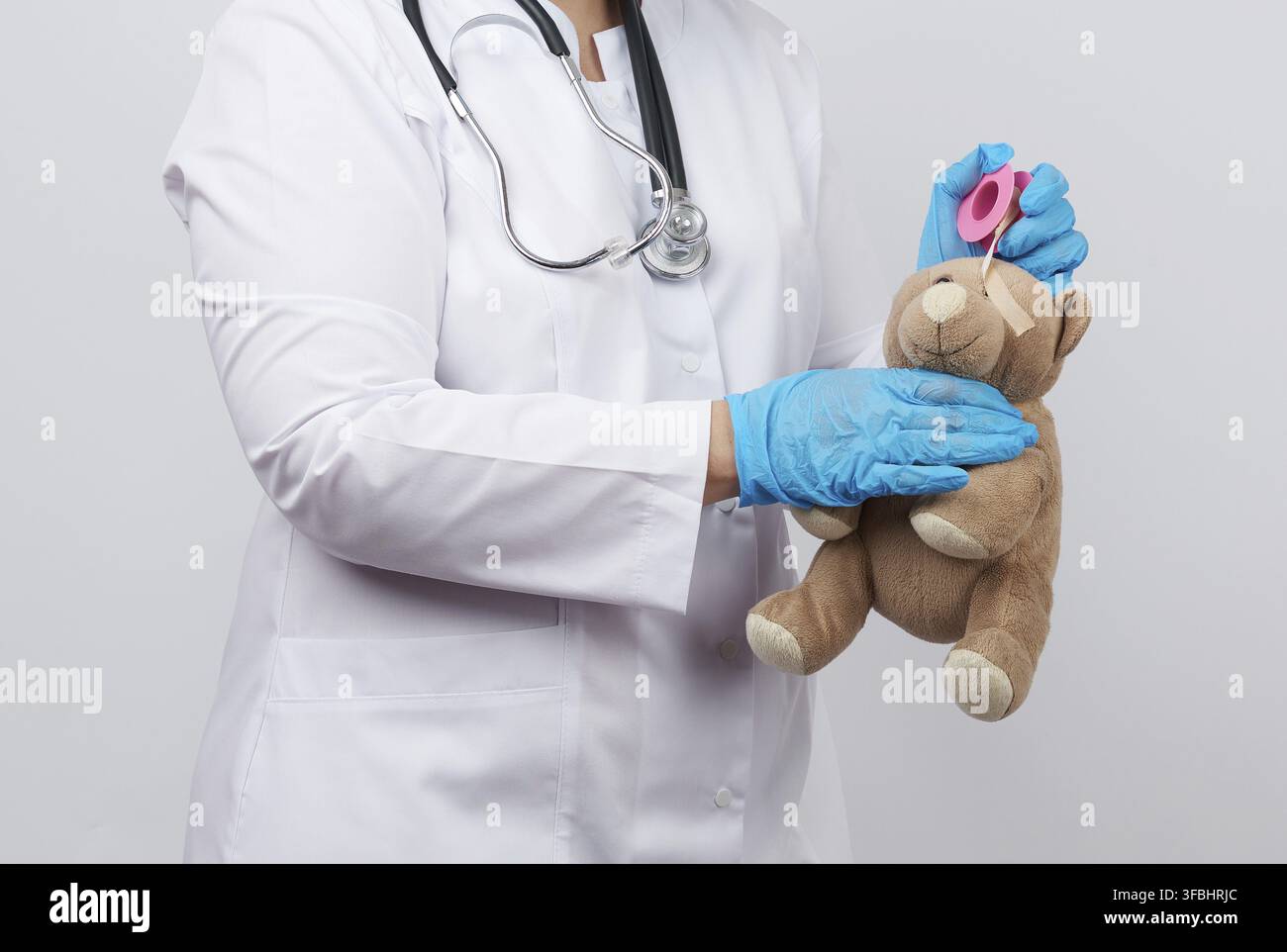 medico femminile in un camice bianco e guanti di lattice blu tiene un orsacchiotto marrone senza occhio, medico avvolge la testa con cerotto adesivo Foto Stock